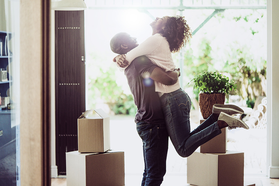 A photo of happy man lifting woman in new house. Side view of loving and excited couple are in casuals. They are with cardboard boxes at entrance.